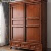 Three-quarter angled lifestyle view of the Casa Furnishing sliding door wooden wardrobe positioned next to a bright window with elegant curtains, showcasing the rich wood grain of the solid wood almirah and its three base drawers in a cozy bedroom setting.